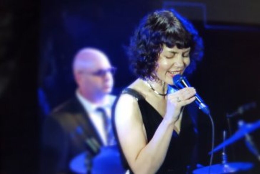 Woman with dark hair, wearing v-neck, sleeveless black dress, holding a microphone on a stage and singing. Musician partially visible behind left.