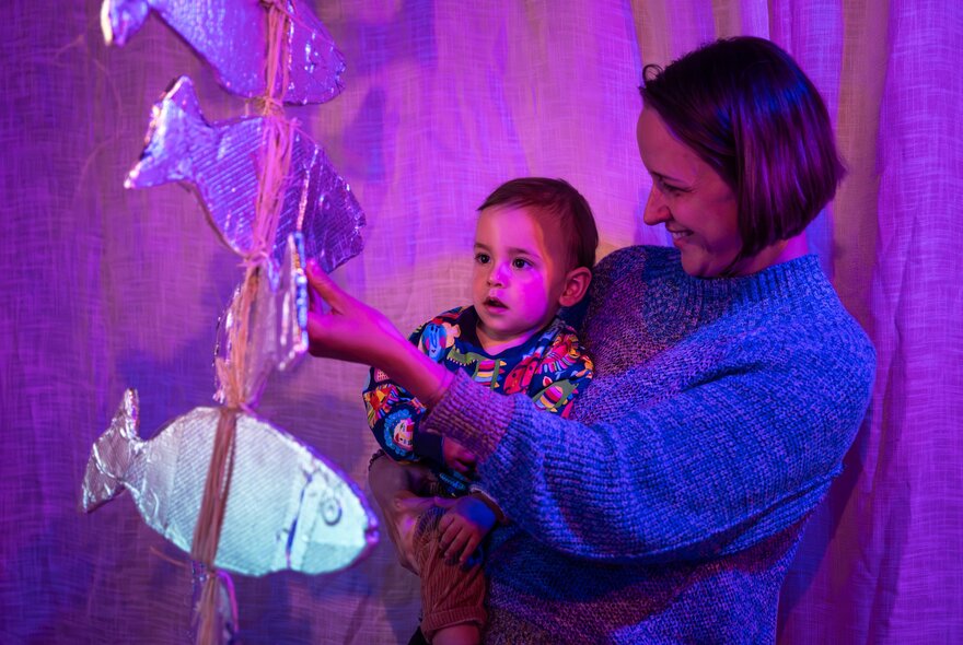 A smiling woman holds a toddler in her arms as the woman reaches out to touch a large hanging fish mobile; part of an interactive installation at ArtPlay.