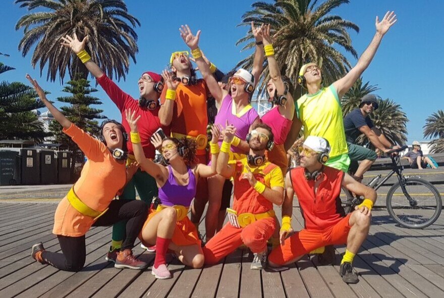 Guru Dudu Silent Disco Walking Tour with participants wearing headphones and bright retro style 1980 clothing, striking a group pose on a boardwalk with palm trees in the background. 