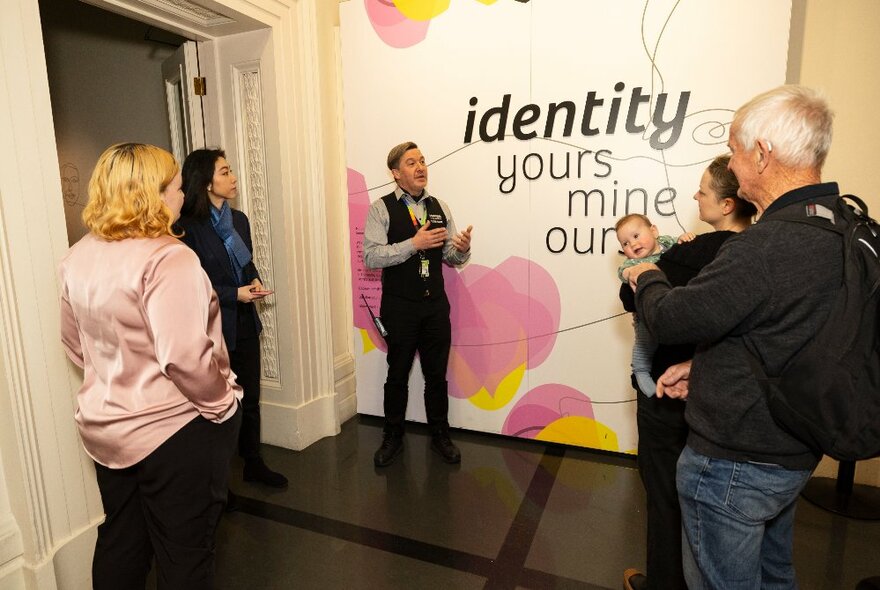 People on a tour at the Immigration Museum all listening to a speaker standing in front of a wall mural saying 'identity, yours, mine, ours'.