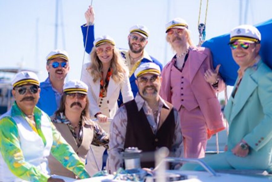 Members of Yacht Rock Revival band posing on a yacht in colourful 1970s-style suits, moustaches and captains hats.