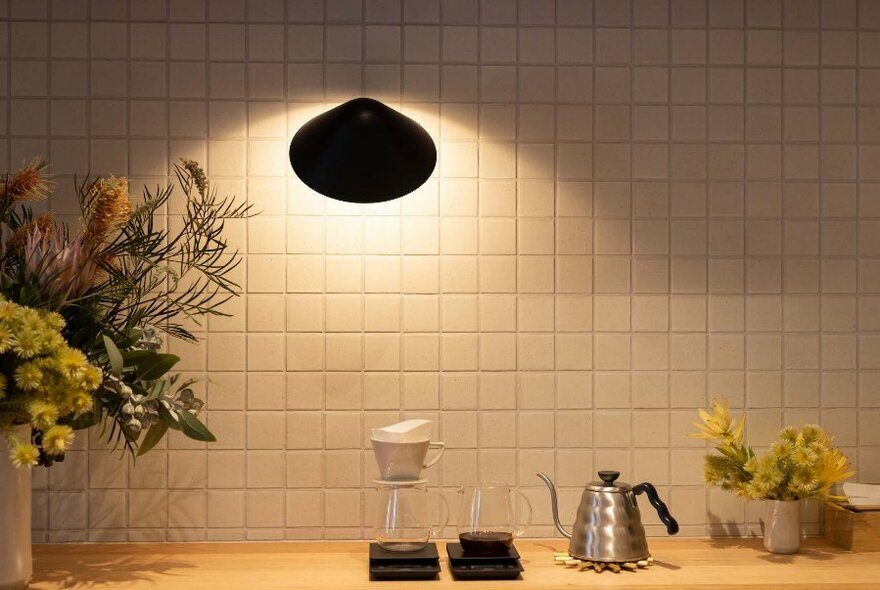 Minimalist white tiled wall, with a spotlight on two glass filter coffee jugs and vases of flowers, on a wooden counter. 