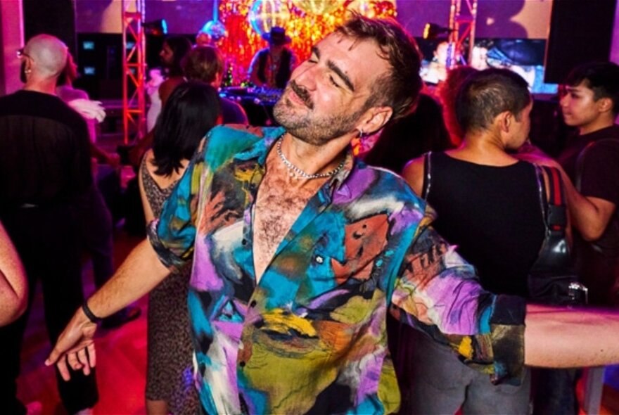 A man wearing a multi-coloured suit and no shirt dances with his eyes closed with people in the background. 