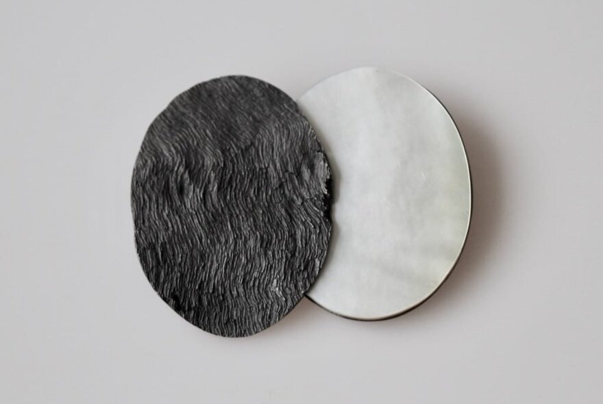 A contemporary piece of jewellery of two overlapping ovals, one dark, the other light, against a white background.