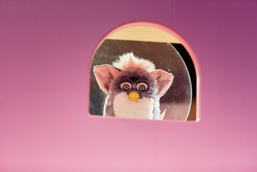 A small, fluffy, brown and white Furby toy with large eyes and ears, peeking out from a pink, arched opening.
