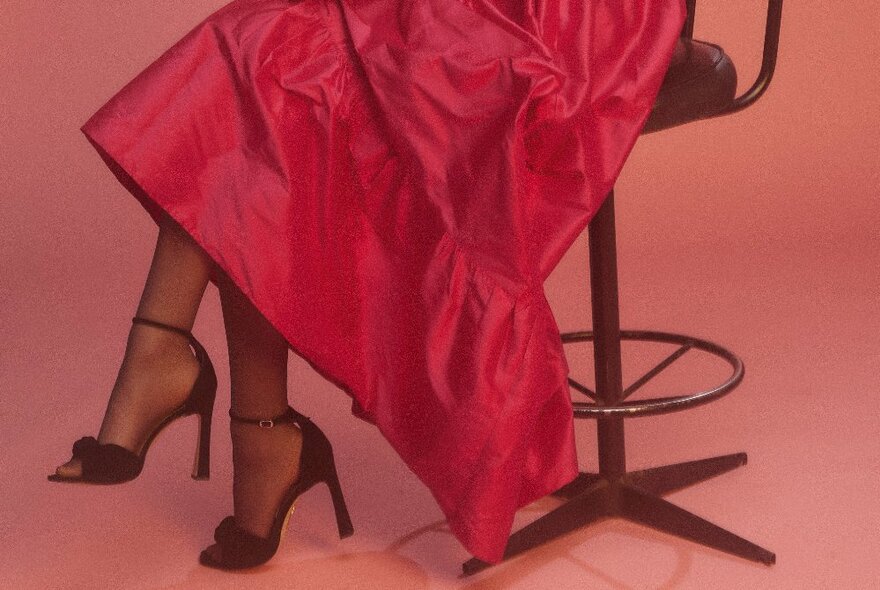 The lower part of a fashion model seated on a stool with her red dress reaching her crossed ankles; she is wearing high heeled black shoes.