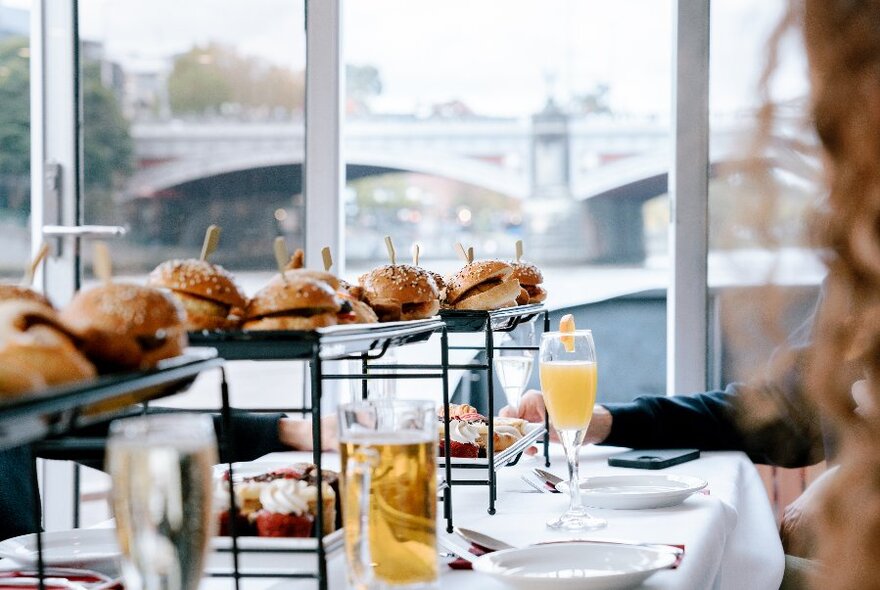 A table laden with burgers and beer, inside a boat passing a view of a bridge in the background.