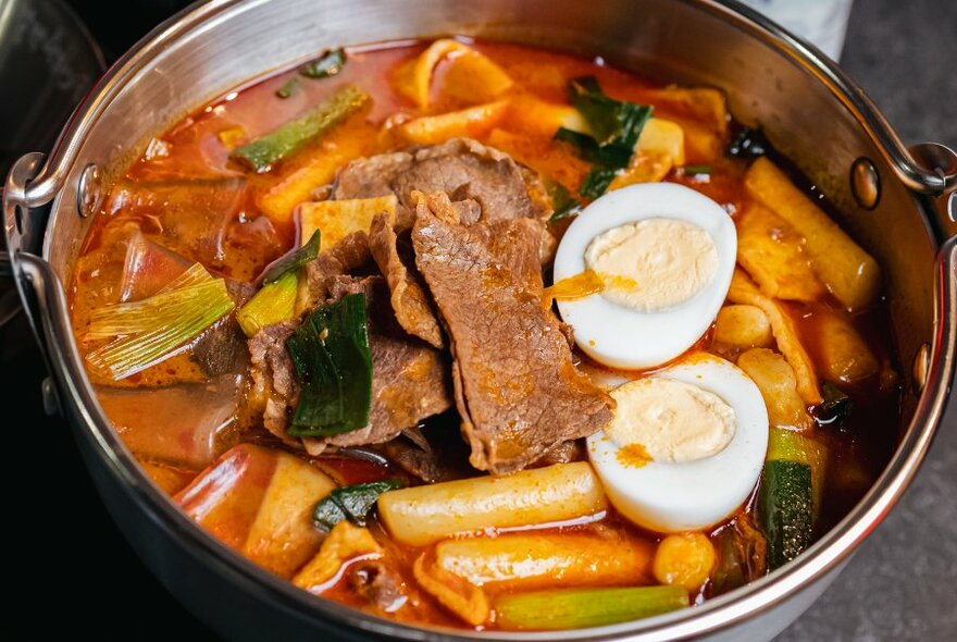 A metal bowl filled with rice cakes, soup and egg.