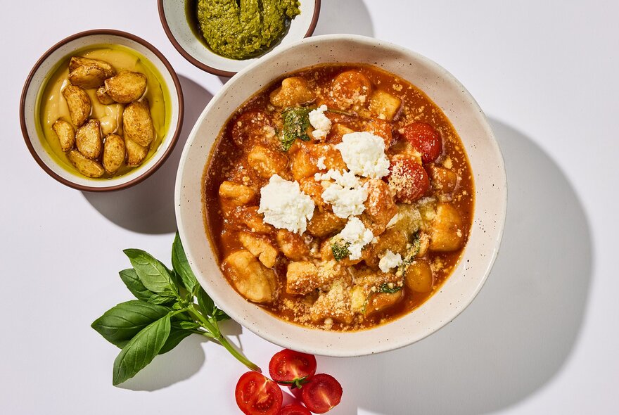 A bowl of tomato gnocchi, seen from above, with fresh cherry tomatoes, basil leaves and other condiments alongside.