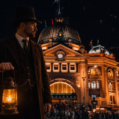 Man holding an old-fashioned lantern outside illuminated Flinders St Station at night, wearing a suit and cap.