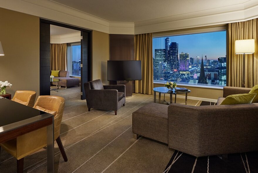 Hotel suite with lounge room and separate bedroom, large picture window at dusk with panoramic city view.