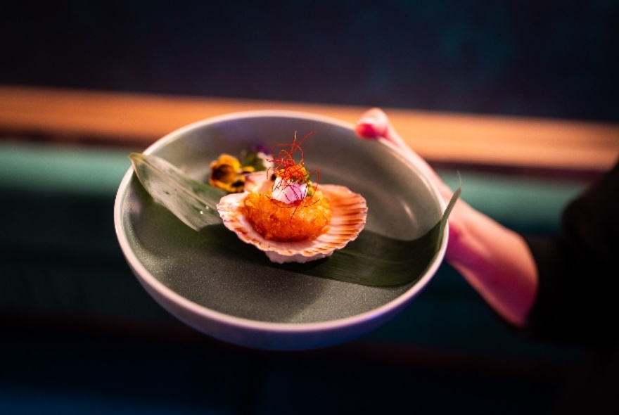 A woman's hand holding a ceramic plate with a cooked scallop sitting on a banana leaf, with edible flowers as garnish.