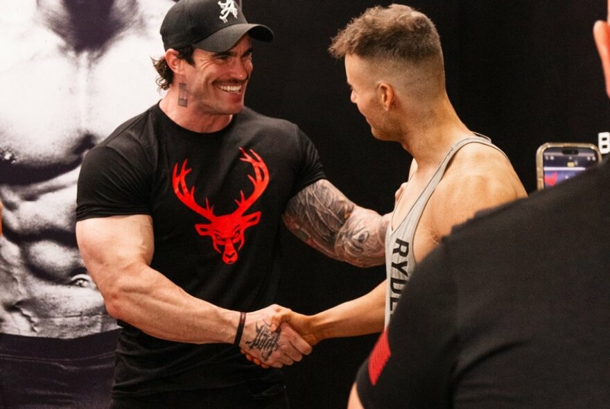 Two men with large muscled biceps smiling shaking hands.