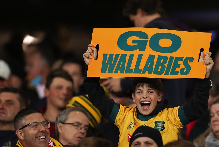 Sports fans in a stadium wearing Wallabies colours, a young boy holding up a yellow sign with green letters that read GO WALLABIES.