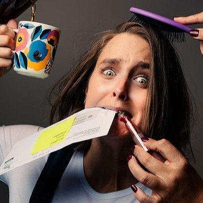 A frazzled woman holds up a mug while coffee is poured into it, someone brushes her hair and someone else puts lipstick on her lips all while she's holding an envelope in her teeth.