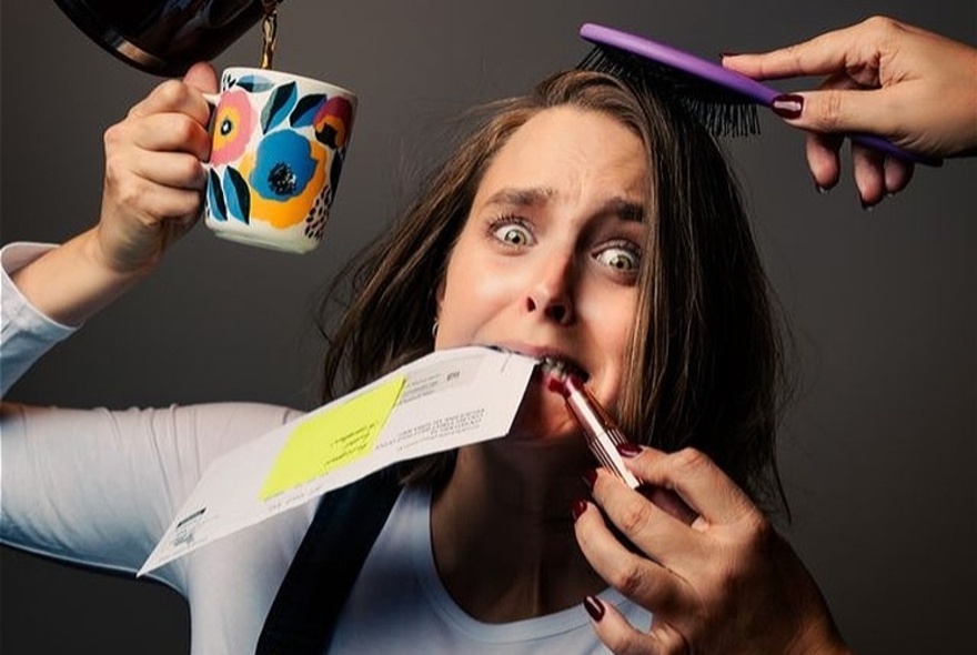 A frazzled woman holds up a mug while coffee is poured into it, someone brushes her hair and someone else puts lipstick on her lips all while she's holding an envelope in her teeth.