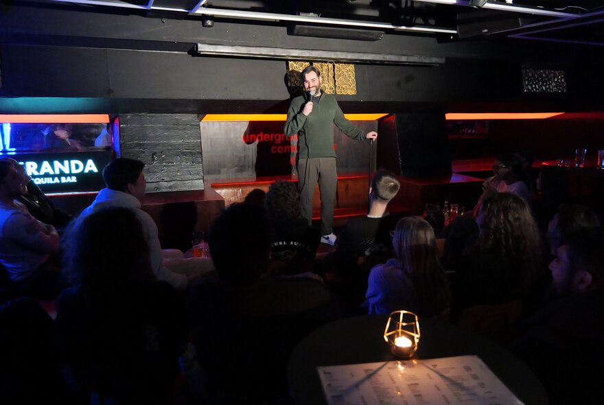 A live stand-up comedy performance in a dimly lit basement venue or club with an audience seated in the foreground.