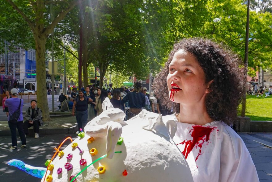 An individual with curly hair wearing a white, textured costume adorned with colourful round elements and fake blood dripping from their mouth, standing outdoors in a city street with people and a park in the background.