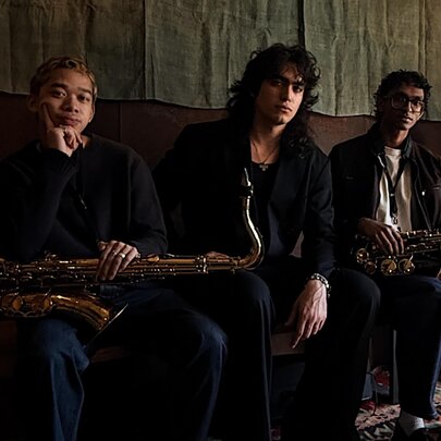 Three piece jazz band, two of the members holding saxophones, seated on a bench seat inside a dimly lit room.
