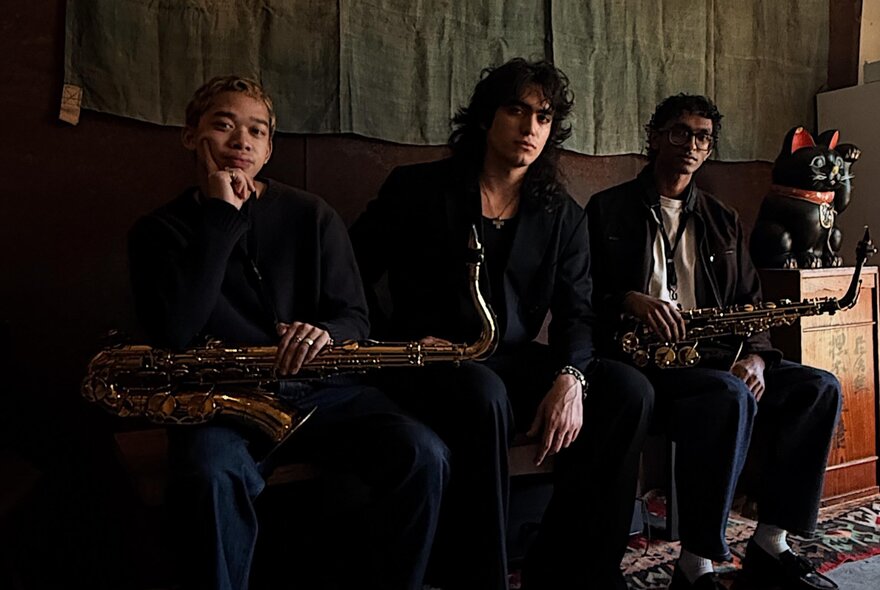 Three piece jazz band, two of the members holding saxophones, seated on a bench seat inside a dimly lit room.