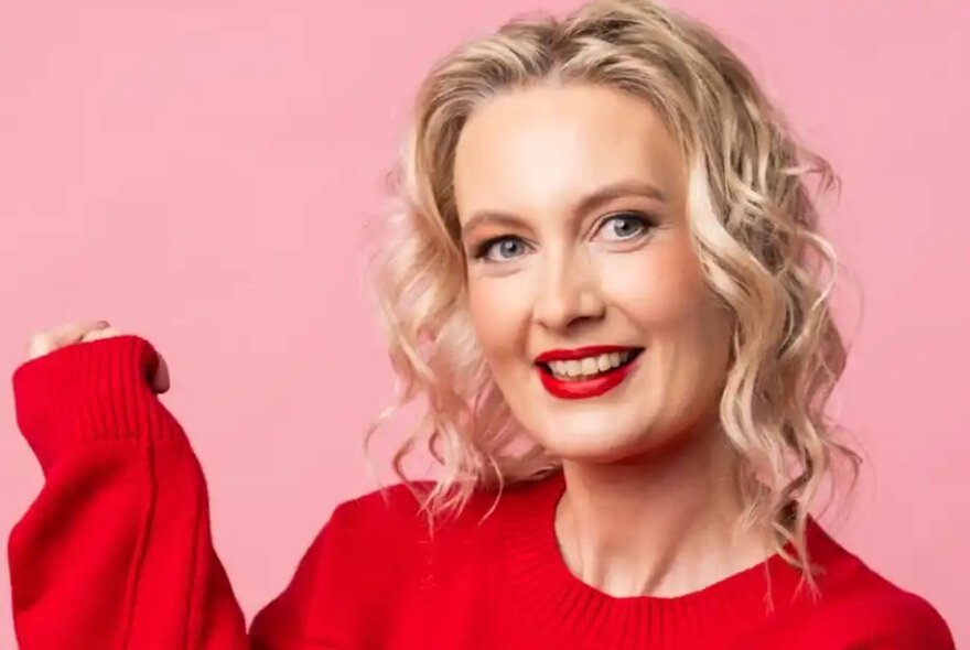Blonde woman wearing a red jumper, holding her arm up, her hand hidden.