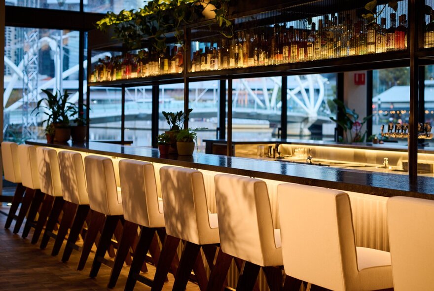 White high bar stools lined up under the bar of 
Crane Bar & Lounge at 1 Hotel Melbourne, with a view through the large windows behind the bar onto the Yarra River.