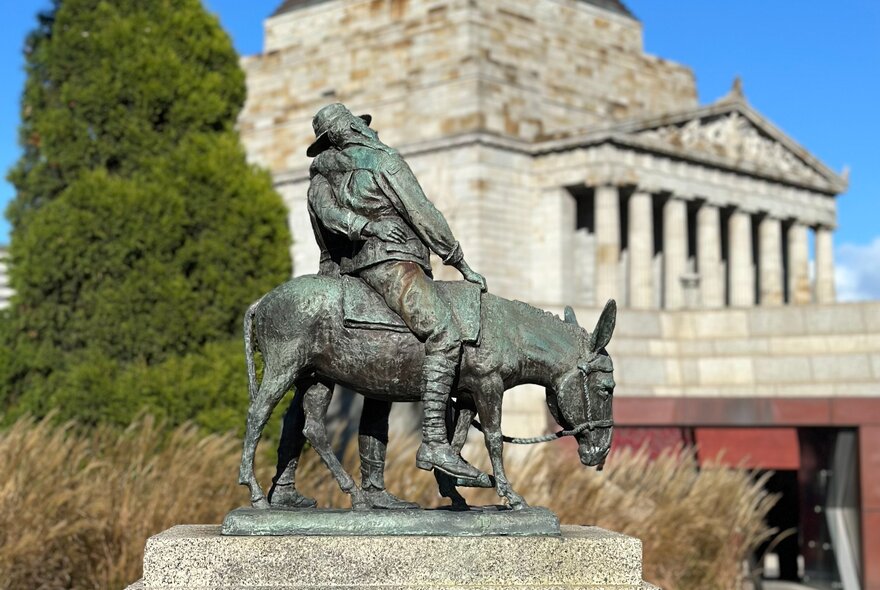 A bronze statue of 'The Man with the Donkey', showing a man leading a donkey carrying a wounded soldier, set against the backdrop of the Shrine of Remembrance. 