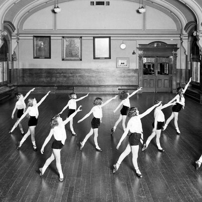 Vintage black and white photograph of a dance class taking place inside the ballroom on the third floor of Melbourne's Flinders Street Station, shows a group of young people in white tops and black shorts performing a dance routine in a large hall with a wooden floor and arched ceiling.