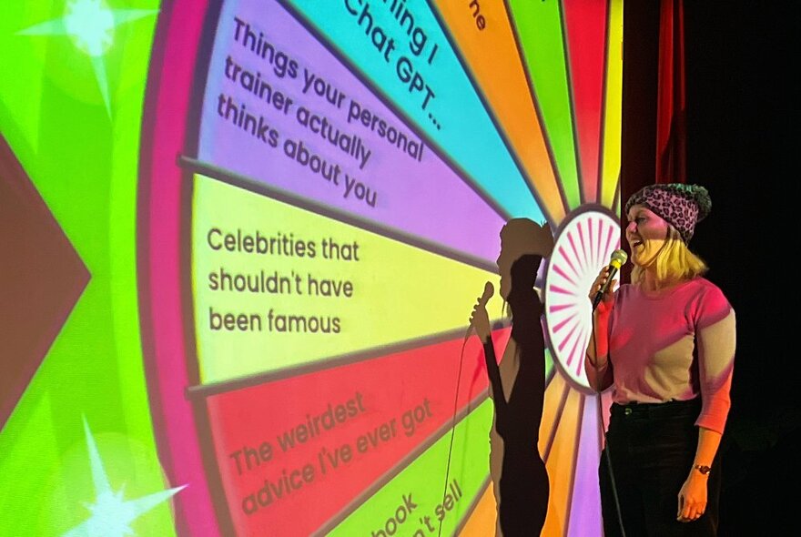 A comedian on stage in front of a projected spinning wheel that has prompt phrases printed in the segments.