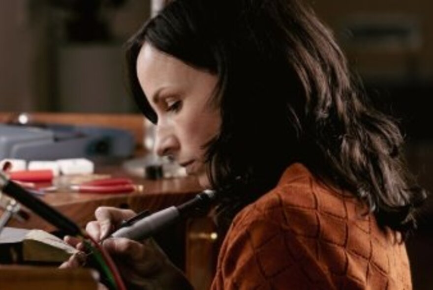 Profile of a woman with long dark hair working with instruments close to her face in a workshop. 