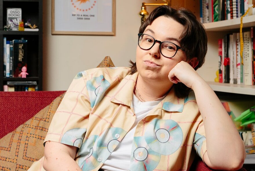 Woman seated on a couch in a living room with bookshelves, her chin on her hand, wearing spectacles with short dark hair.