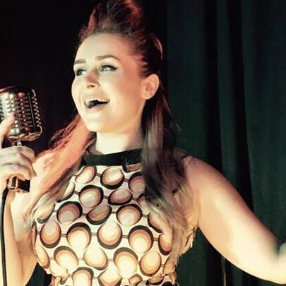 A singer in a 1960s-style dress holds a retro microphone with one arm outstretched.