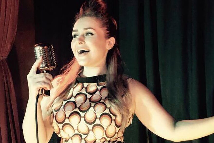 A singer in a 1960s-style dress holds a retro microphone with one arm outstretched.