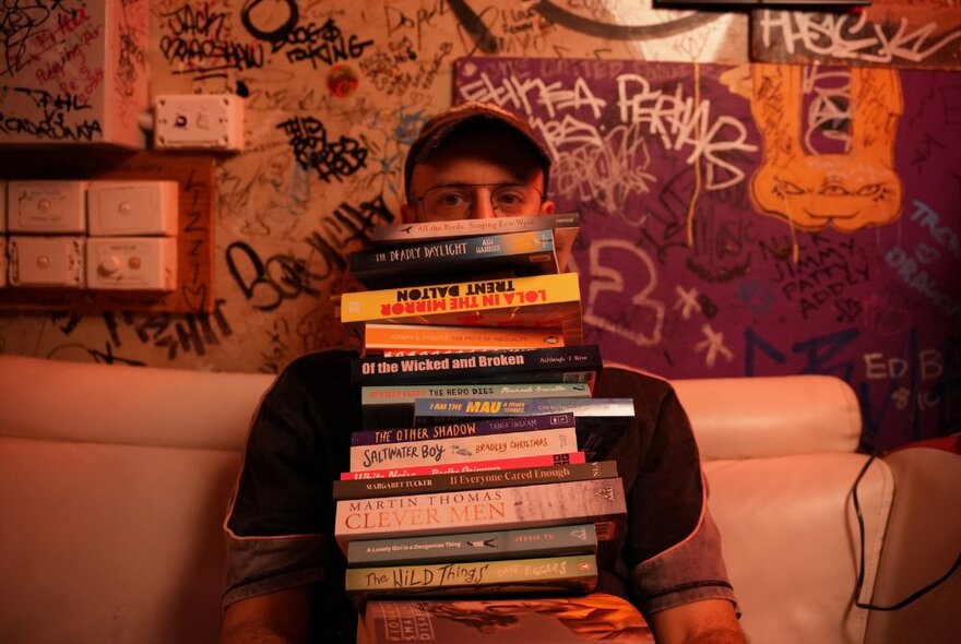 A person holding a large stack of books in front of their face, posed against a graffiti-covered wall.