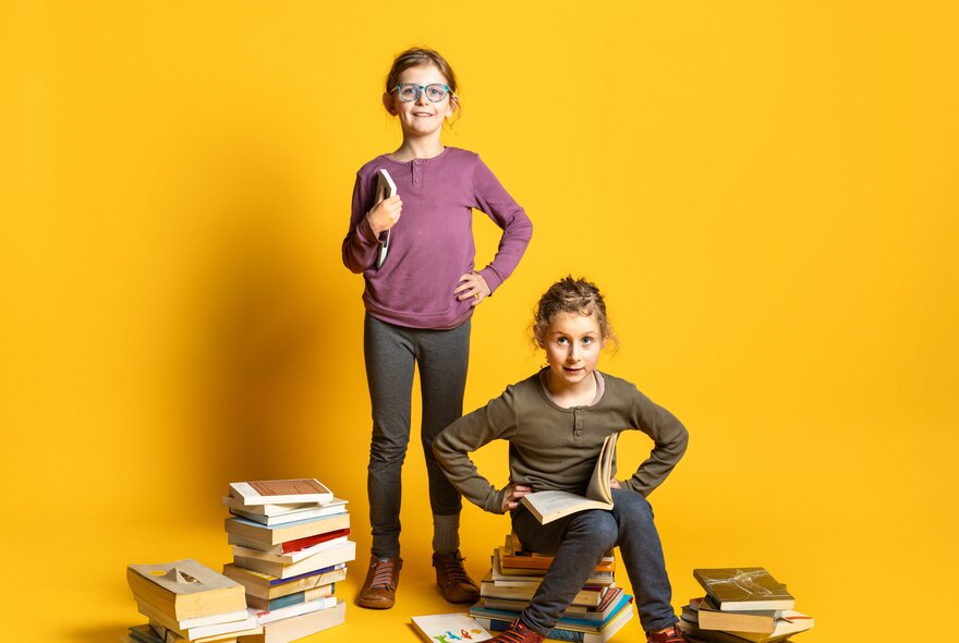 Two girls in a yellow room, one standing and one seated, next to piles of books