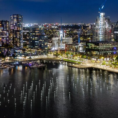 Night aerial view of the Melbourne Docklands skyline with the calm waters of Victoria Harbour and a public art installation of illuminated buoys in rows in the foreground. 