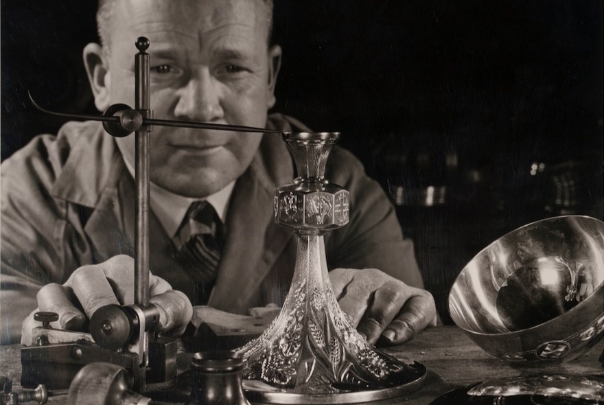 A black and white portrait of a man looking at an elaborate glass and silver carafe from the level of the tabletop.