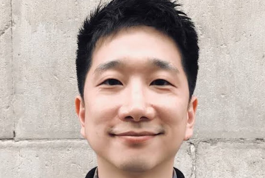 Writer Anton Hur with short black hair, standing against white brick wall.