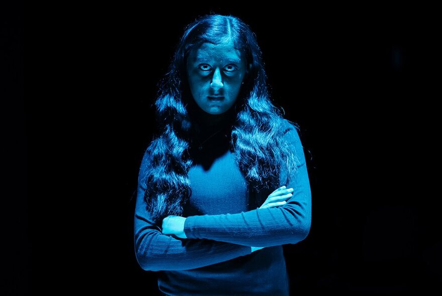 A teenager with a sullen expression on their face, their arms crossed in front of their chest standing against a dark background and bathed in a blue light.