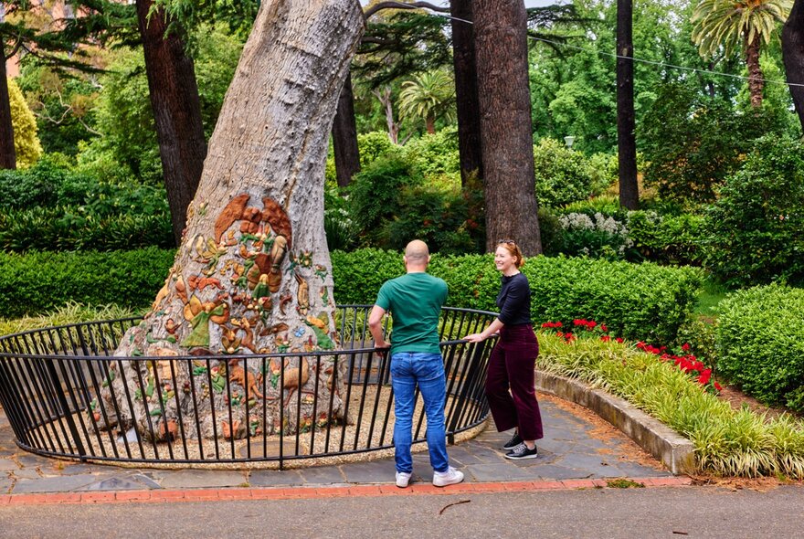Two people standing near a large, textured tree trunk surrounded by a black metal protective fence in a garden.