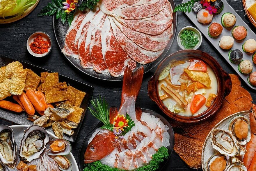 Overhead view of plates of raw food including seafood and very thinly sliced beef, waiting to be cooked in a hot pot.