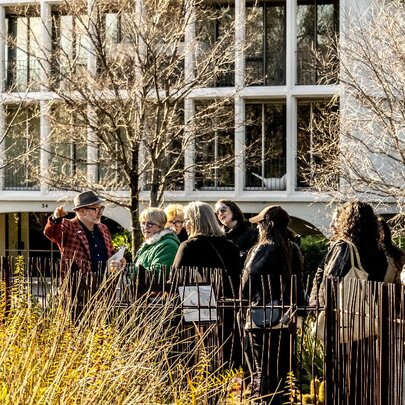 A small group of people on a tour on a pathway with yellow grass on one side and a large building on the other. 