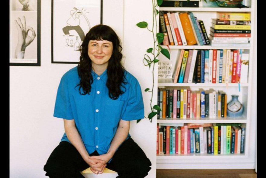 Author Sinéad Stubbins, sitting on a stool in her home next to a colourful bookshelf, black and white framed artwork behind her on the white wall.