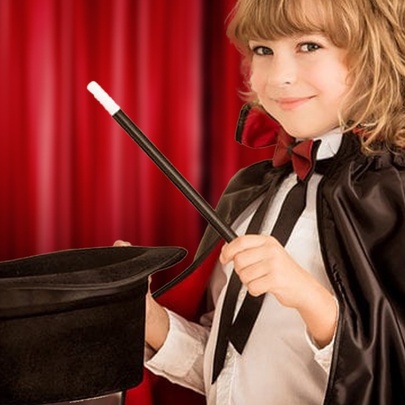 A child wearing a magician's cape, holding a wand and top hat in front of red curtains.