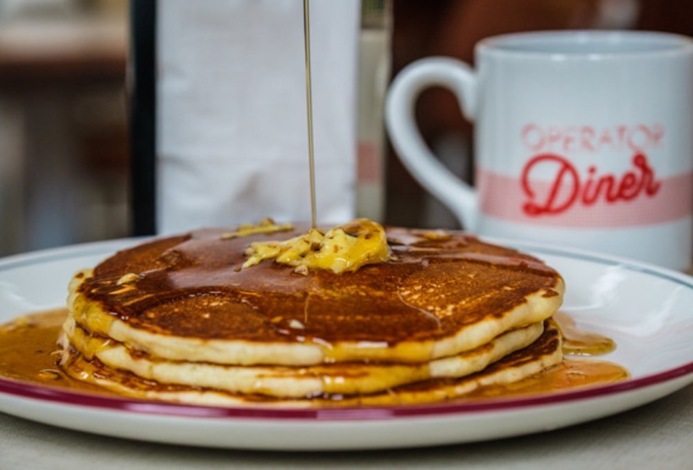 A stack of pancakes with butter and maple syrup being poured on top.