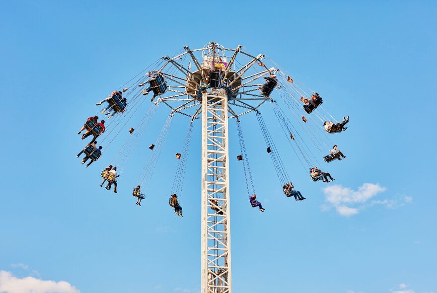 A carnival ride showing people strapped into high chairs and being swung around the top of a pole, set against a blue sky.