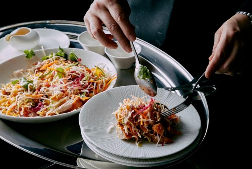 Hand using a spoon and fork to dish food onto a smaller plate from a larger bowl.