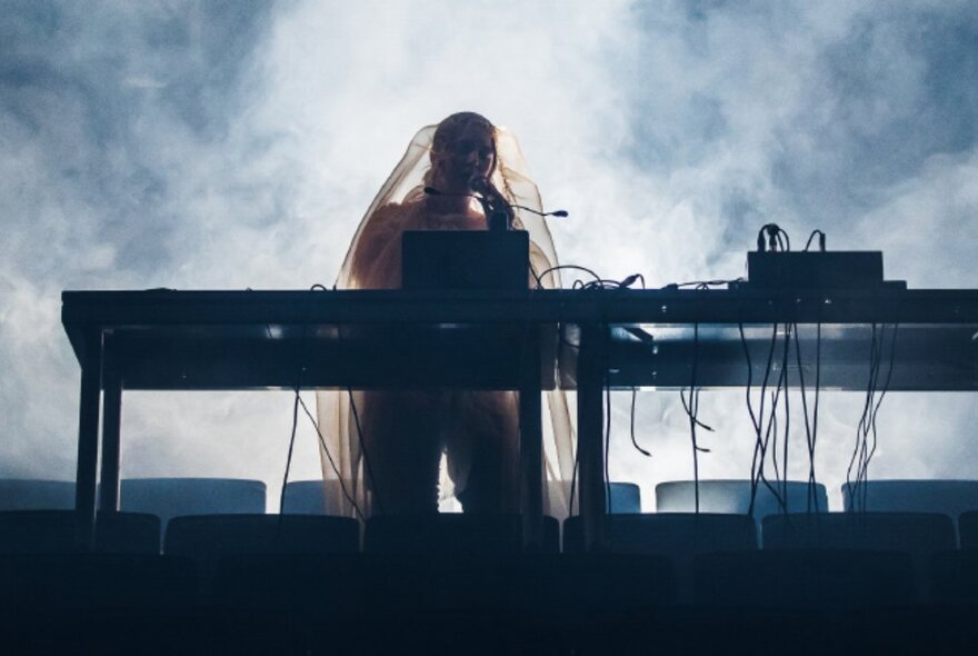 A musician silhouetted and performing on stage behind an electronic keyboard and mixing panel, a smoke effect haze surrounding them.