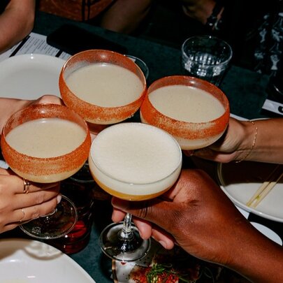 Four cocktail glasses being held together by four separate individuals' hands, over a black table.