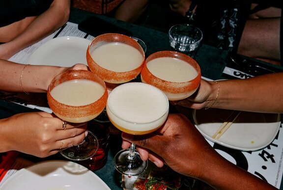 Four cocktail glasses being held together by four separate individuals' hands, over a black table.